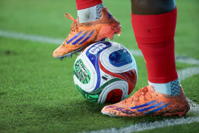 Bota de fútbol naranja en contacto con un balón blanco, mientras un jugador en posición de reposo muestra signos de uso en el campo.
