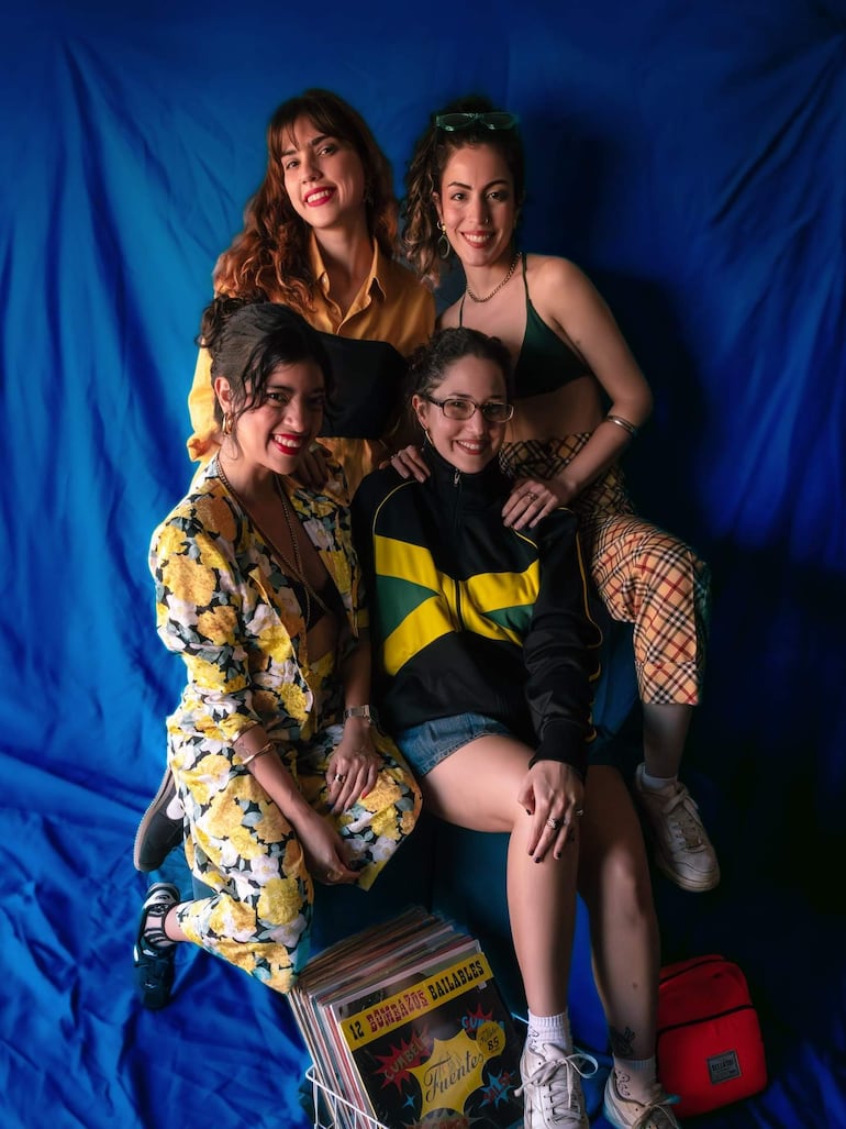 Cuatro mujeres sonrientes posan en un estudio con fondo azul, una con blusa amarilla y otra con top de bañador, disfrutando el momento.