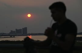 Un hombre aplaca el intenso calor tomando tereré en la Costanera de Asunción. De fondo se ve la silueta de los nuevos edificios del gobierno en la Bahía de Asunción, durante una puesta de sol.