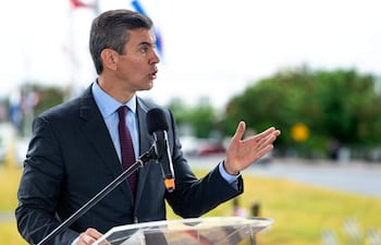 Santiago Peña Santi en traje oscuro gesticulando frente a un atril, con banderas y un paisaje verde de fondo.