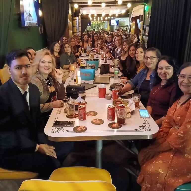 Grupo de personas sonrientes en un ambiente festivo, con comida y bebida sobre una mesa larga, decoraciones luminosas en el fondo.
