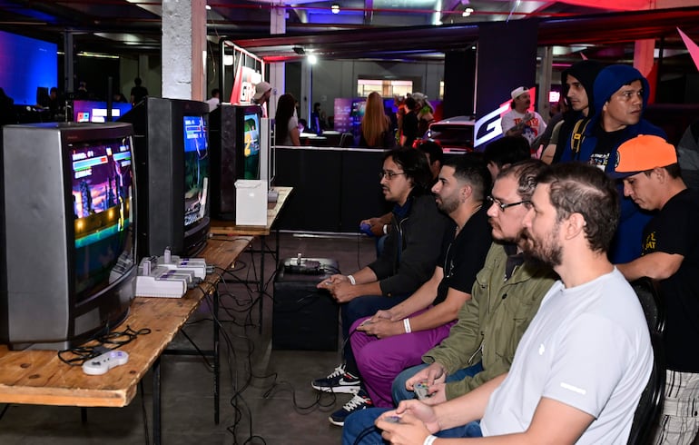 Hombres concentrados jugando videojuegos, con controles en mano y televisores antiguos en fondo de luces de colores.
