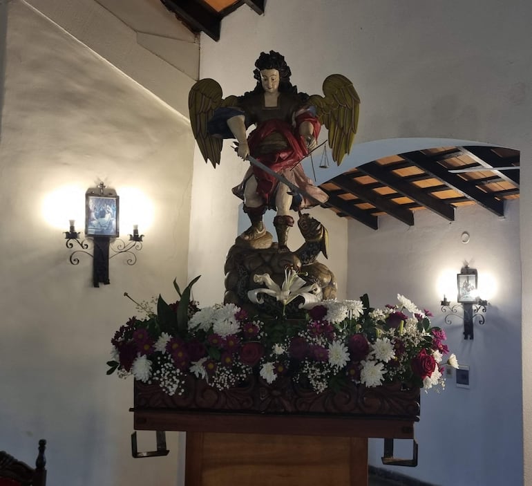 Escultura del Arcángel San Miguel con alas doradas y espada, en altar con arreglos florales en un ambiente solemne.