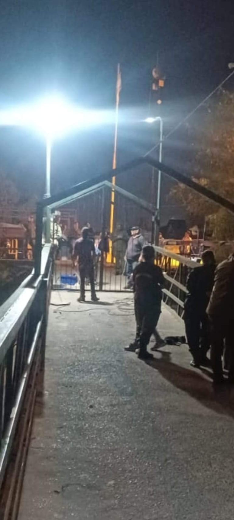 Varias personas dialogan en un control de seguridad iluminado, mientras un individuo supervisa la entrada.