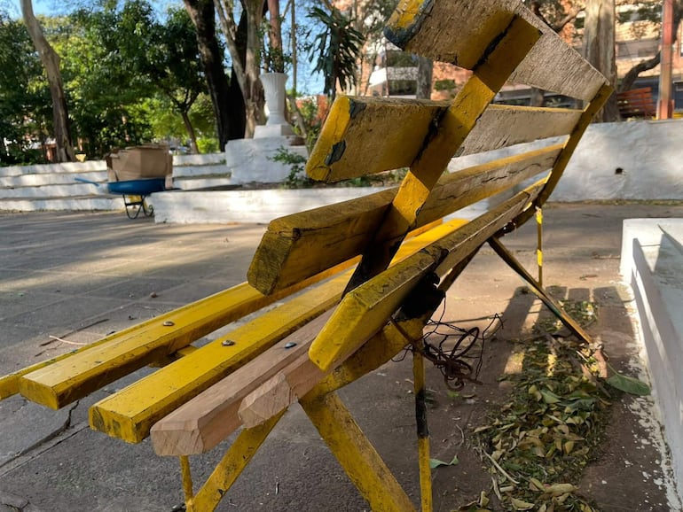 Banco de parque amarillo deteriorado, rodeado de vegetación, en la plaza Batallón 40 de Asunción.