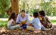 Una mujer adulta sonríe y lee un cuento a dos niñas y dos niñas sentados a su alrededor, sobre hojas secas, a la sombra de un árbol.