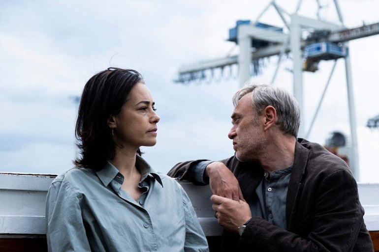 Mujer con cabello oscuro y camisa clara escucha atentamente a hombre de cabello canoso con chaqueta oscura, en ambiente de puerto nublado.