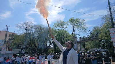 Hombre con traje blanco y gafas oscuras sostiene objeto en llamas, rodeado de manifestantes con carteles y banderas en un entorno urbano.
