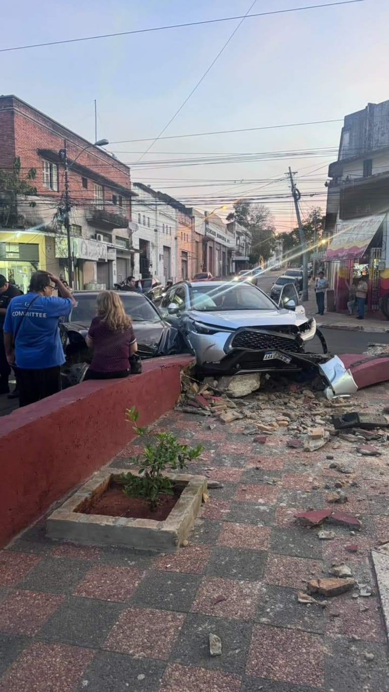 Hombre en camiseta azul analiza el accidente, mientras dos mujeres, una de blusa negra y otra junto al coche, observan la escena.