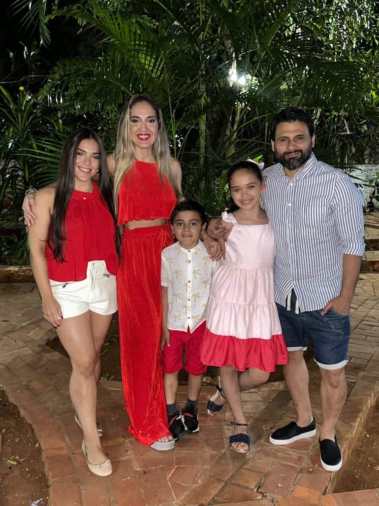 Mujer de pie con cabello lacio y atuendo rojo, niño en centro con camiseta blanca y pantalones rojos, rodeados de vegetación.