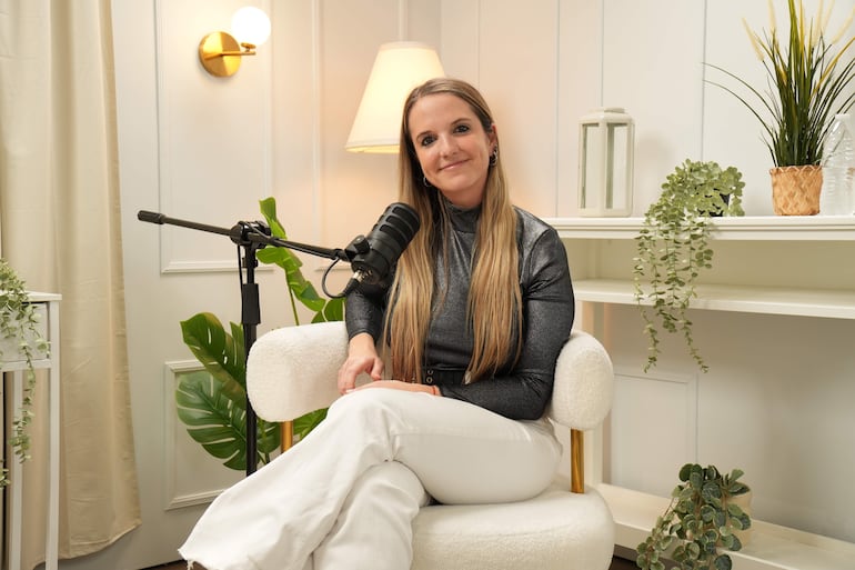 Mujer sonriente con blusa negra y pantalones blancos sentada en un sillón frente a un micrófono en un estudio decorado con plantas.