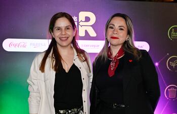 Dos mujeres sonrientes en un evento, una con top negro y pantalones de flores, la otra en traje negro con pañuelo rojo.