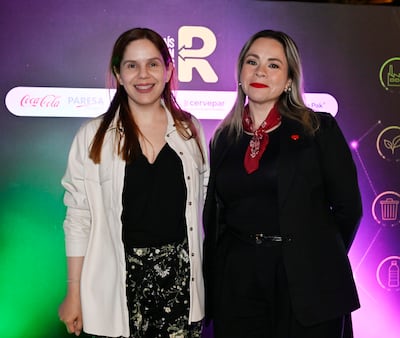Dos mujeres sonrientes en un evento, una con top negro y pantalones de flores, la otra en traje negro con pañuelo rojo.