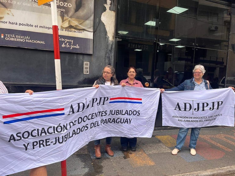 Cuatro mujeres sostienen banderas con el texto ADJPJP, vestidas casualmente, mostrando seriedad en una manifestación frente al MEC.