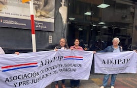 Cuatro mujeres sostienen banderas con el texto ADJPJP, vestidas casualmente, mostrando seriedad en una manifestación frente al MEC.