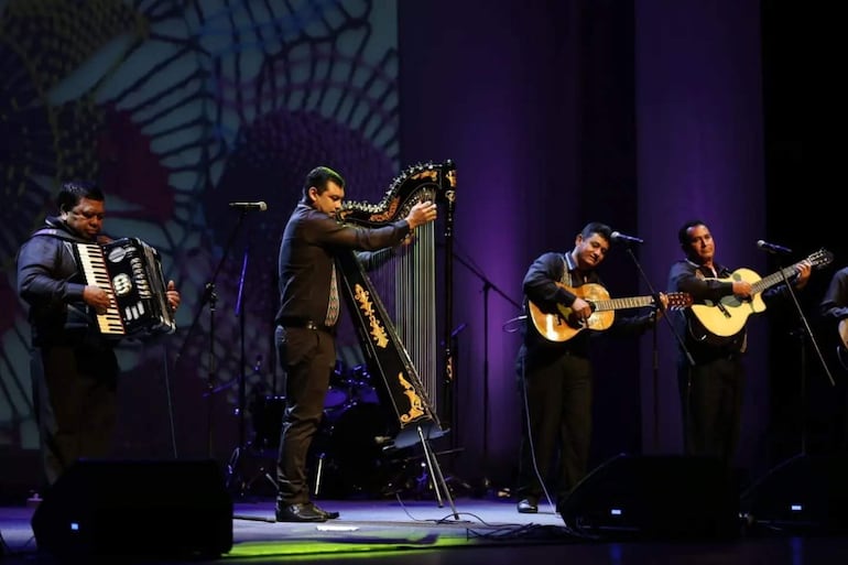 Cuatro músicos vestidos con camisas oscuras, concentrados en sus instrumentos: acordeón, arpa y guitarras, en un escenario iluminado con luces púrpuras.