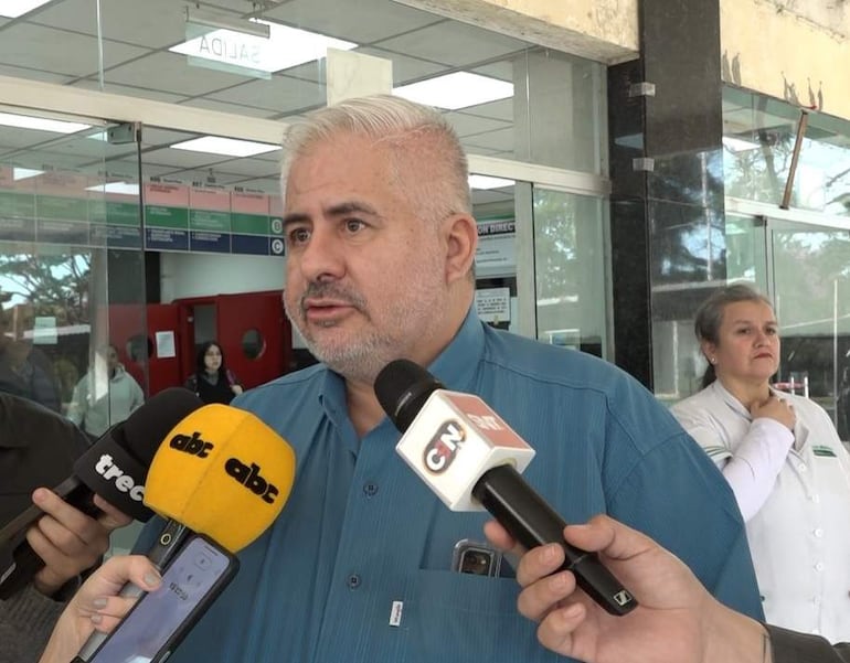 Alvarenga con camisa azul sostiene un micrófono, rodeado de otros medios y una mujer en uniforme blanco, en ambiente de atención a la prensa.