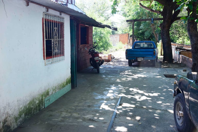 Patio con una motocicleta negra y una camioneta azul, rodeado de árboles y una pared blanca descuidada.