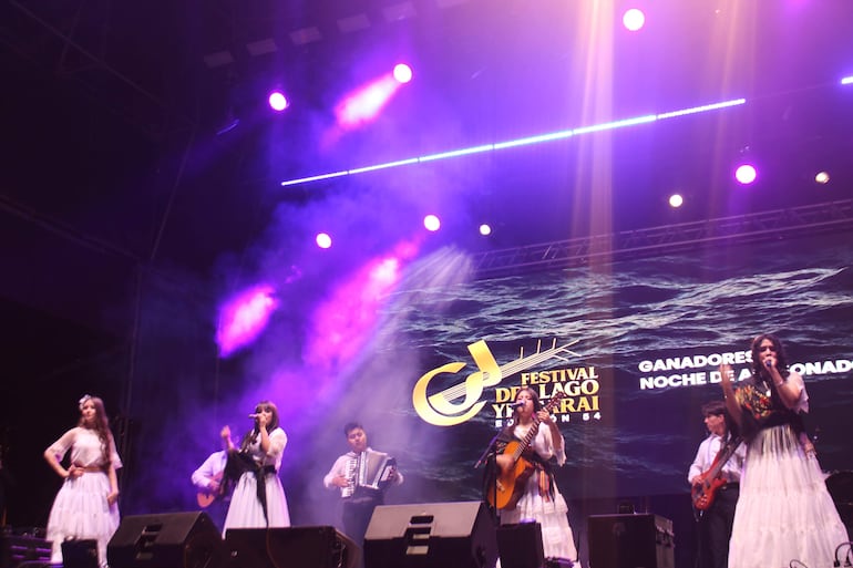 Vocalistas con trajes tradicionales cantan en el escenario colorido del festival, acompañadas por guitarra y acordeón.