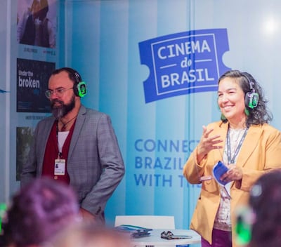 Mujer con chaqueta amarilla habla sonriente, mientras hombre de saco gris escucha atentamente en evento de cine.