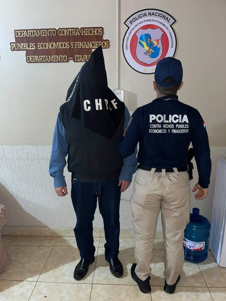 Hombre con capucha a un lado de oficial de policía en entorno del departamento de hechos punibles económicos y financieros.