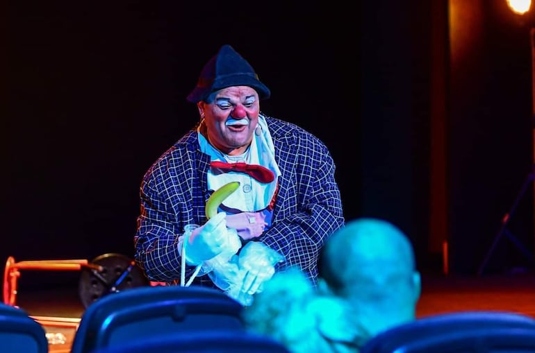 Clown caracterizado con maquillaje blanco y nariz roja, sostiene una banana mientras el público observa con atención en un ambiente iluminado.