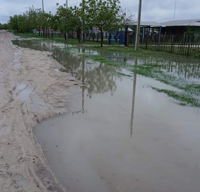 Escenario rural con agua estancada, árboles a la izquierda y edificaciones difusas en el fondo, reflejando inundación.