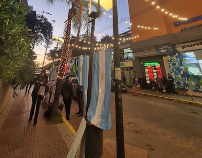 Mujer revisando su teléfono entre otras personas, con banderas argentinas y luces decorativas en un entorno urbano.