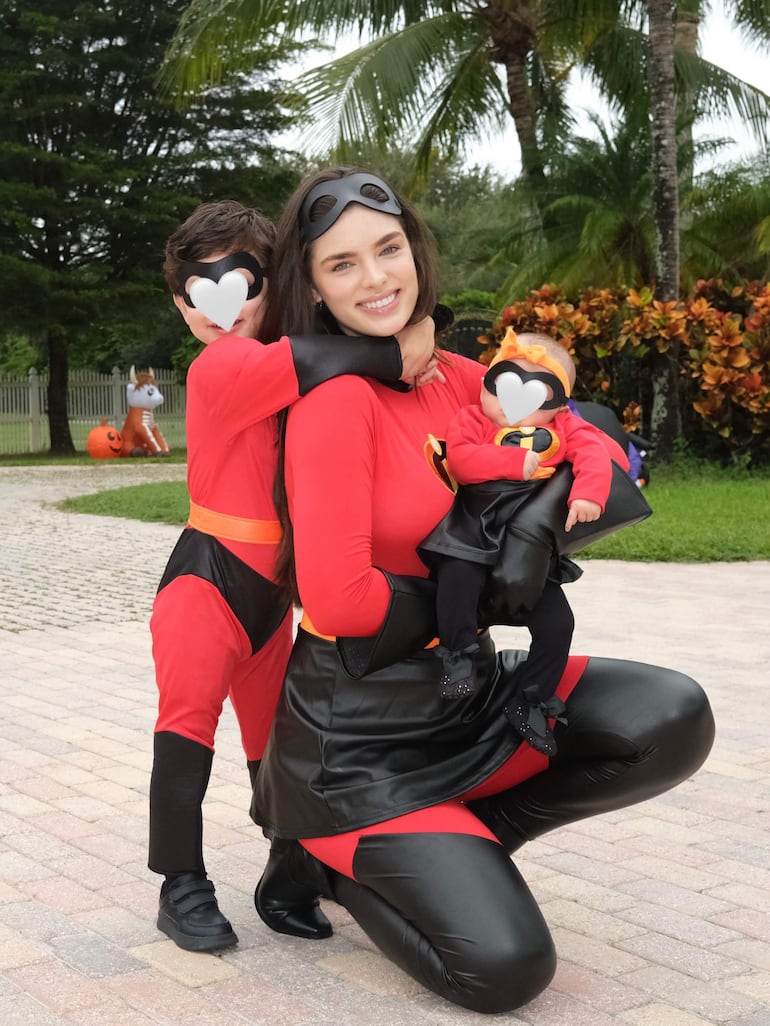 Nadia Ferreira sonríe, vestida de superhéroe rojo, con un niño y un bebé en disfraces similares, rodeados de árboles en un parque.