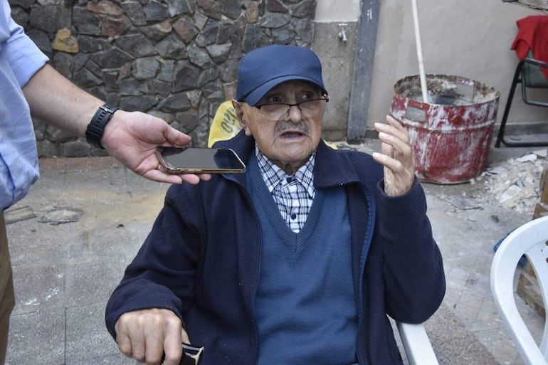 Hombre mayor con abrigo y gorro, habla con un periodista que sostiene un móvil, en un ambiente sencillo al aire libre.