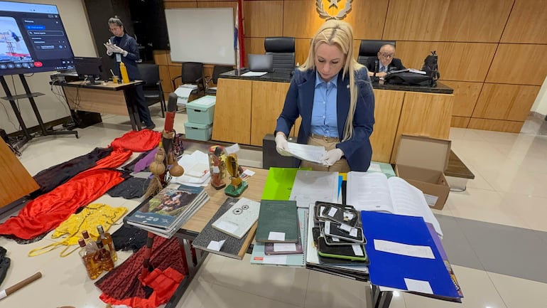 Mujer rubia con camisa azul y guantes revisa documentos; hombre mayor con lentes observa en fondo.