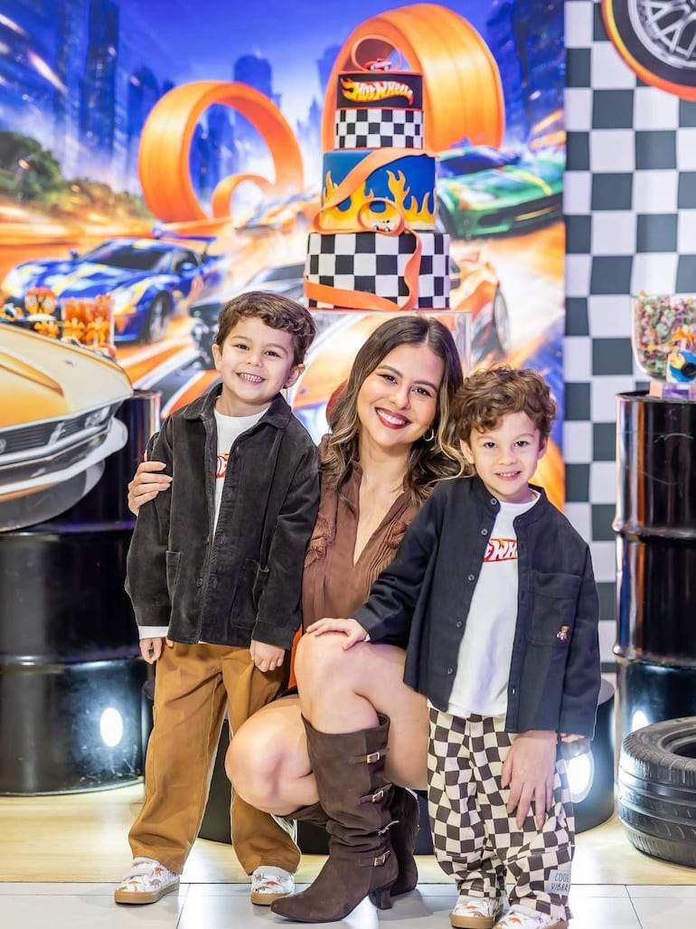 Mujer sonriente con blusa marrón y dos niños en prendas informales, rodeados de decoración festiva y una torta temática de 'Hot Wheels'.