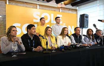 Soledad Núñez, vestida con chaqueta blanca, habla en conferencia rodeada de otros políticos, con cartel amarillo de fondo.