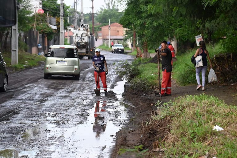 Hombre con camisa azul oscuro y pantalones rojos sostiene una pala en calle con barro; mujer con bolsa camina a un lado.