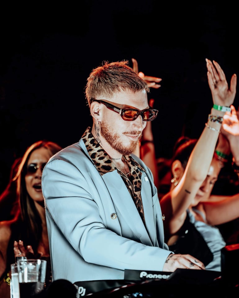 Hugel con chaqueta azul pálido y gafas oscuras, mientras el público aplaude y levanta las manos en un ambiente vibrante.