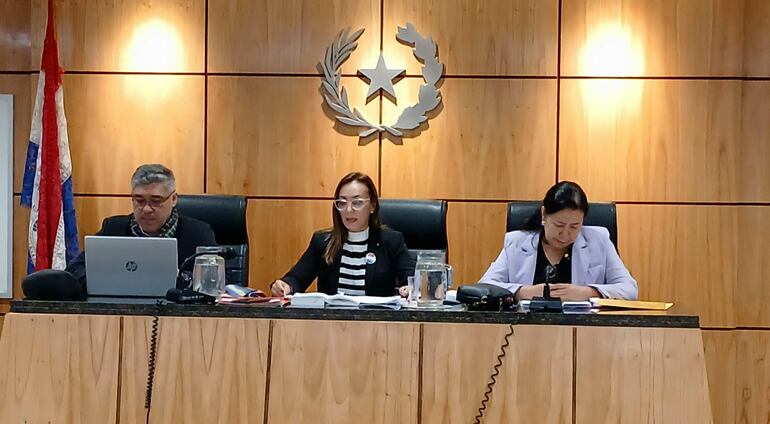 Mujer con chaqueta negra y rayas blancas presenta documentos, mientras dos adultos la observan en un ambiente formal con bandera paraguaya.