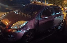 Automóvil púrpura dañado en su parte delantera, estacionado en una vía pública húmeda tras la lluvia.