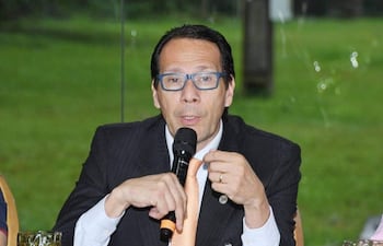 Hombre con camisa blanca y corbata naranja sostiene micrófono, luz natural entra por ventanas, ambiente de reunión formal.