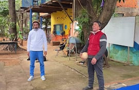 Hombre con camisa blanca y pantalones claros escucha mientras otro, con suéter rojo, sostiene una pelota en un patio colorido.