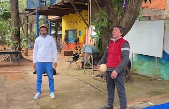 Hombre con camisa blanca y pantalones claros escucha mientras otro, con suéter rojo, sostiene una pelota en un patio colorido.