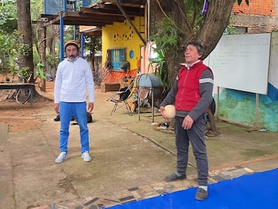 Hombre con camisa blanca y pantalones claros escucha mientras otro, con suéter rojo, sostiene una pelota en un patio colorido.