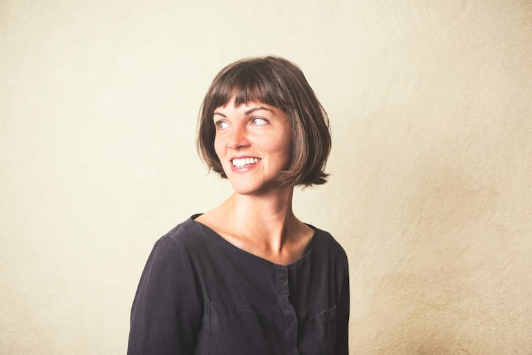 Mujer de perfil con cabello corto y blusa oscura sonríe; fondo claro resalta su expresión alegre.