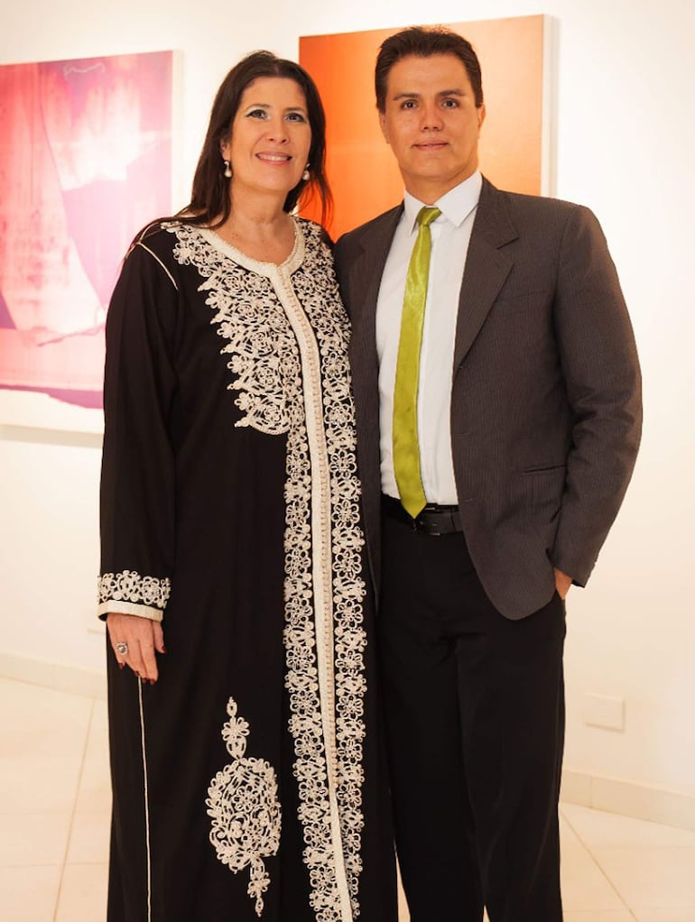 Mujer con túnica negra y detalles blancos sonríe junto a hombre en traje gris y corbata verde en galería iluminada con arte colorido.