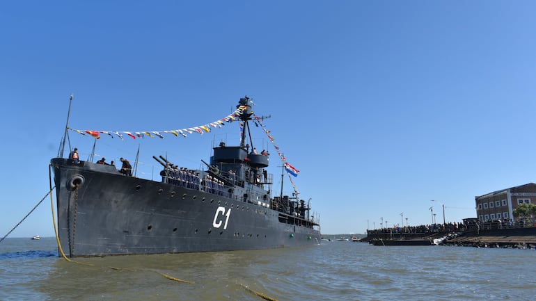 Barco militar C1 con oficiales en uniforme azul, rodeado de banderas, mientras la multitud observa desde el muelle.