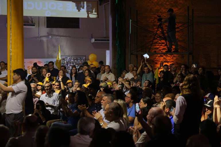 Ricardo Flecha en camiseta blanca sostiene un teléfono, mientras asistentes aplauden en un ambiente festivo con globos amarillos.