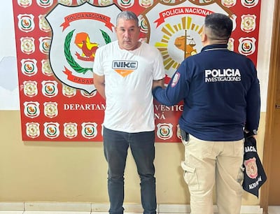 Hombre con camiseta blanca y pantalones oscuros, acompañado por oficial de policía en chaqueta, en ambiente formal de comisaría.