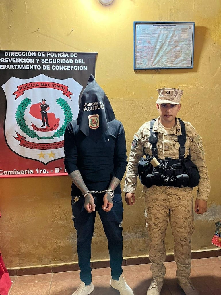 Hombre con pasamontañas negro y esposas, custodiado por policía en uniforme camuflado, en ambiente de comisaría.