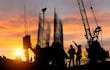 Dos trabajadores en casco y guantes, uno señala hacia arriba mientras el otro escucha, en un sitio de construcción al atardecer.