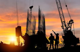 Dos trabajadores en casco y guantes, uno señala hacia arriba mientras el otro escucha, en un sitio de construcción al atardecer.