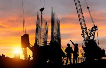 Dos trabajadores en casco y guantes, uno señala hacia arriba mientras el otro escucha, en un sitio de construcción al atardecer.